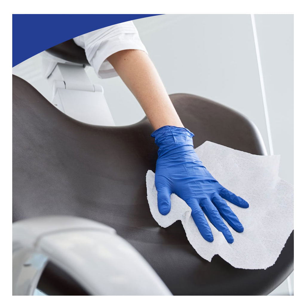 A close-up of a healthcare worker wearing a blue nitrile glove using a white disinfectant wipe to clean a dark brown leather medical examination chair