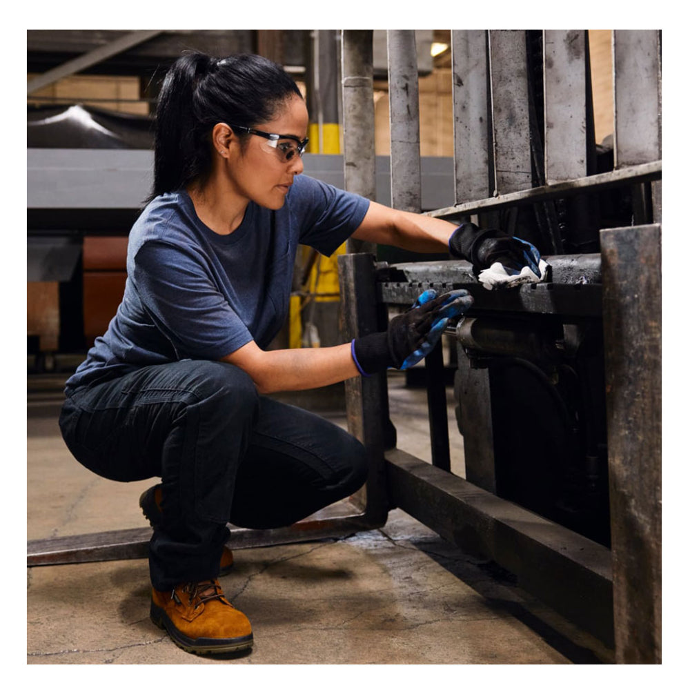A female technician wearing safety glasses and protective work gloves squats down to wipe heavy grease off a forklift assembly using a white WypAll X70 industrial cloth