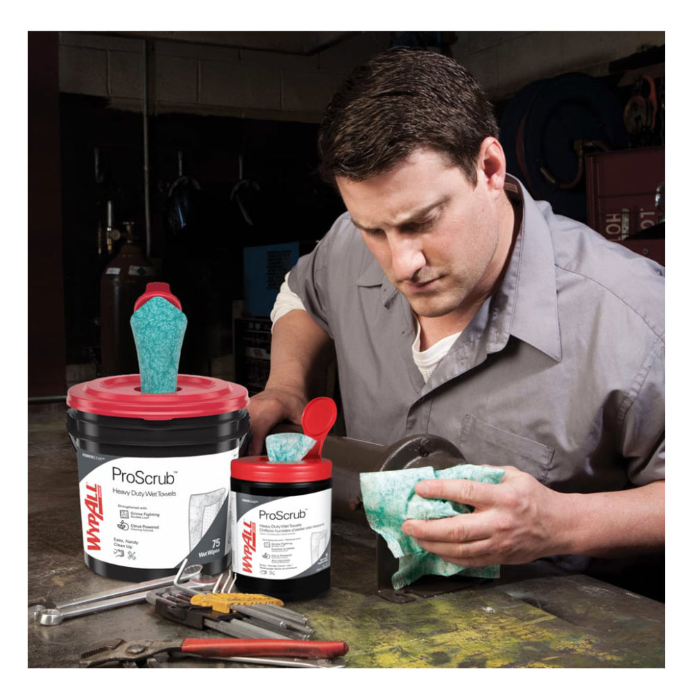 A mechanic in a garage workshop using a green textured WypAll ProScrub heavy-duty wet towel to clean machinery next to dispensing tubs and tools