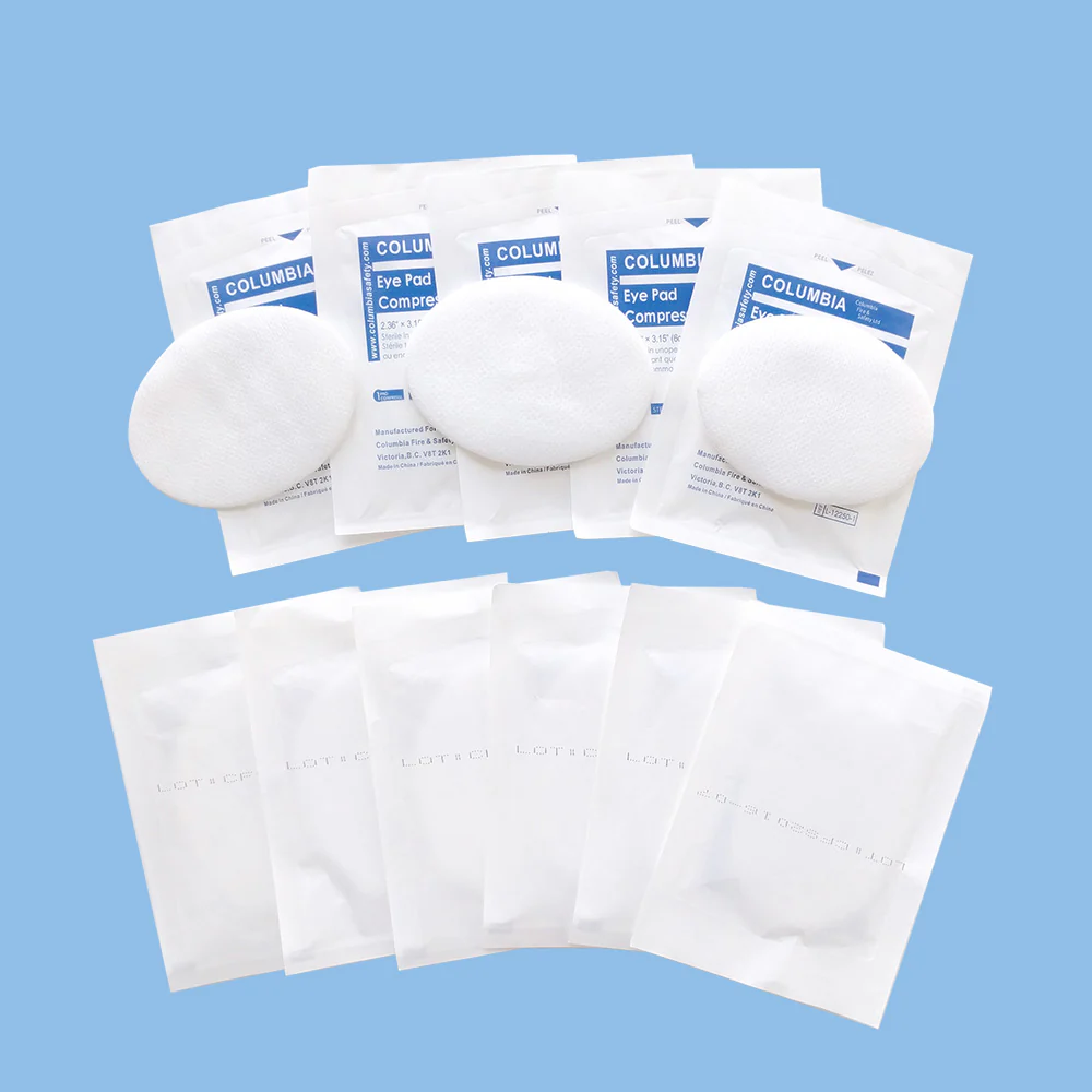 A bulk arrangement of Columbia sterile eye pad compress envelopes laid out on a light blue background with loose oval pads placed on top of the packaging.
