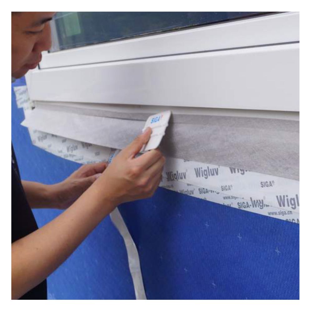 A contractor using a SIGA spreader tool to apply Fentrim 230 grey window flashing tape to a window frame over blue vapor barrier for an airtight seal.