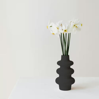 A modern segmented ceramic vase finished in a matte flat black primer coat holding white flowers on a clean white surface.