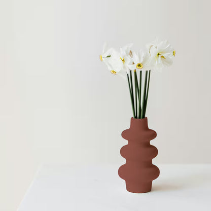 A modern segmented ceramic vase painted in a matte flat red finish holding white flowers on a clean white tabletop.