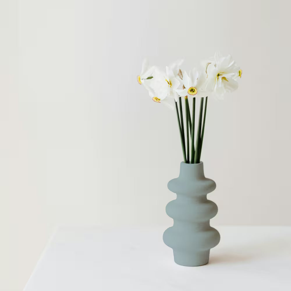 A modern textured ceramic vase painted in a matte grey finish holding white daffodil flowers on a white table.