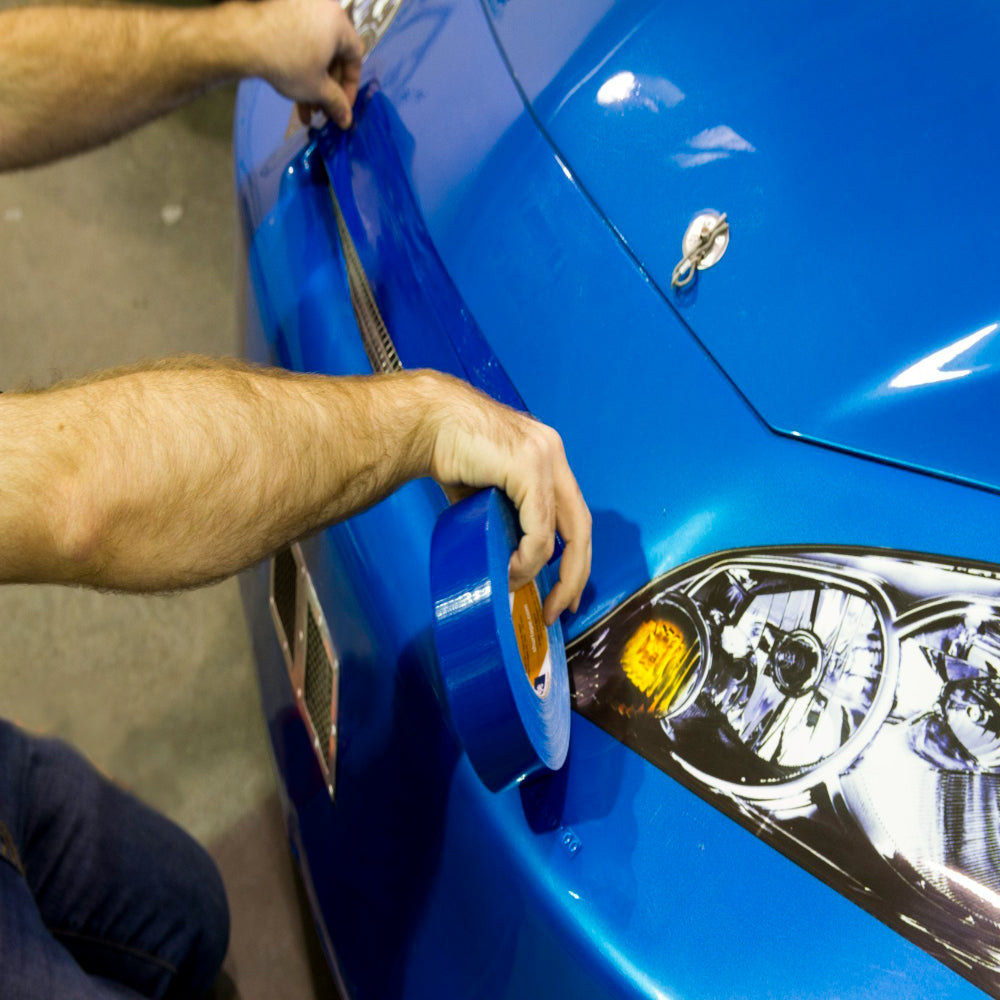 Close-up of a technician applying high-adhesion blue Shurtape duct tape to a blue racing vehicle body for temporary repairs