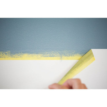 A hand peeling away yellow FrogTape to reveal a clean, sharp paint line between grey and white sections of a wall.