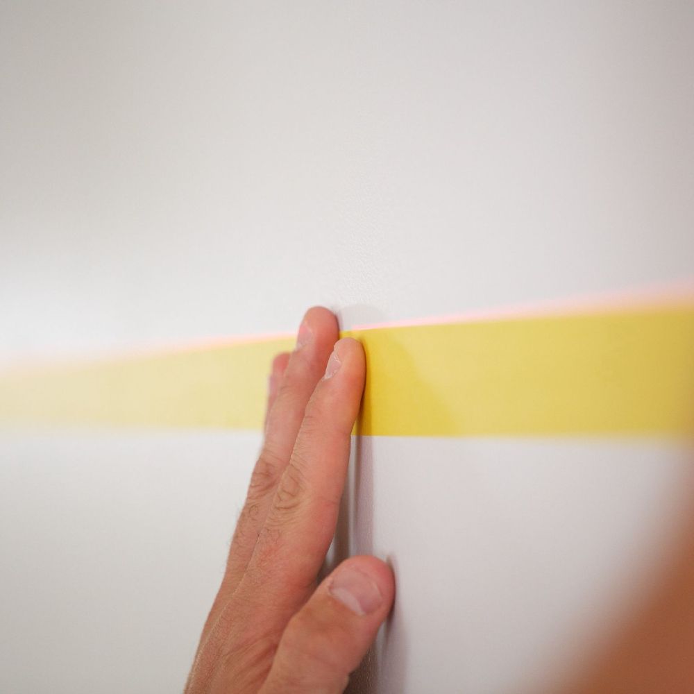 A hand pressing down yellow painter's tape onto a white wall, using a laser level line for perfectly straight placement.