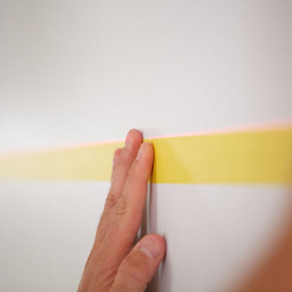 A hand pressing down yellow painter's tape onto a white wall, using a laser level line for perfectly straight placement.