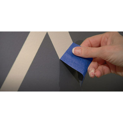 Hand peeling back dark blue painter's tape to reveal sharp geometric paint lines on a wall.