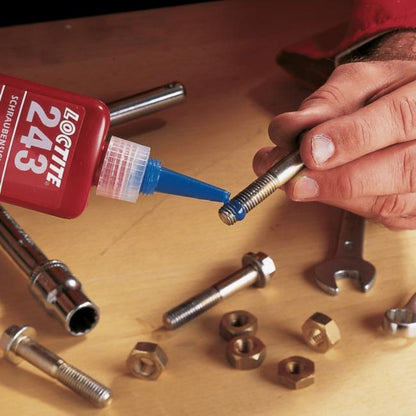 Hands applying Loctite 243 blue medium strength threadlocker to metal bolt threads on a workbench surrounded by nuts bolts and wrenches showing correct application technique