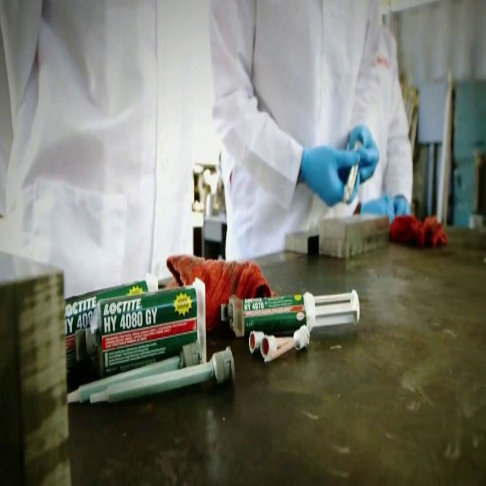 Lab technicians working with Henkel Loctite HY 4080 GY grey structural hybrid adhesive cartridges and mixing nozzles on a workshop table.