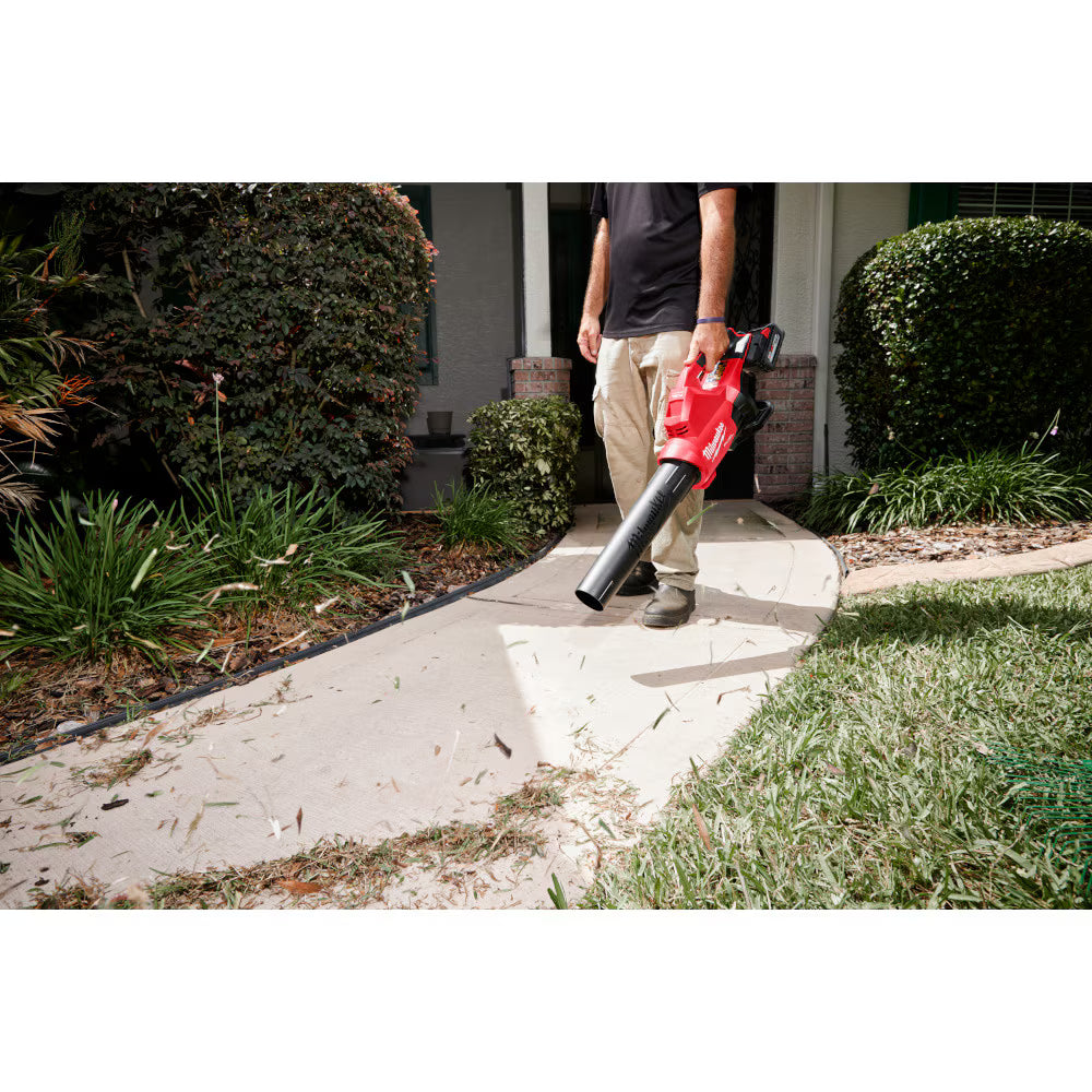A professional landscaper using a Milwaukee M18 FUEL handheld blower to clear leaves and grass clippings from a mulch bed in front of a residential home.