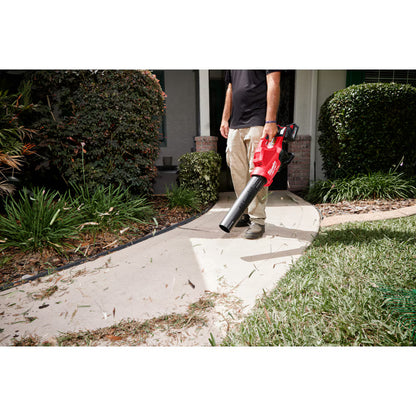 A professional landscaper using a Milwaukee M18 FUEL handheld blower to clear leaves and grass clippings from a mulch bed in front of a residential home.
