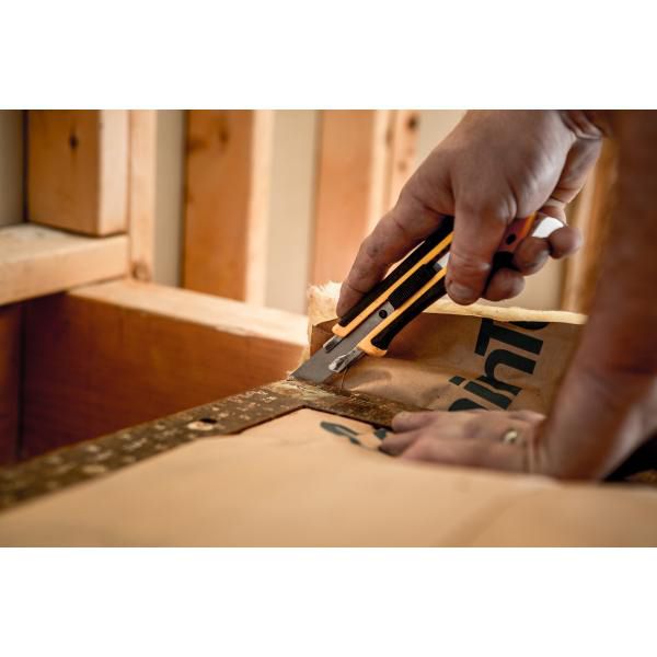 Contractor slicing through heavy kraft backing paper on insulation inside a wooden stud framing wall with an OLFA utility knife
