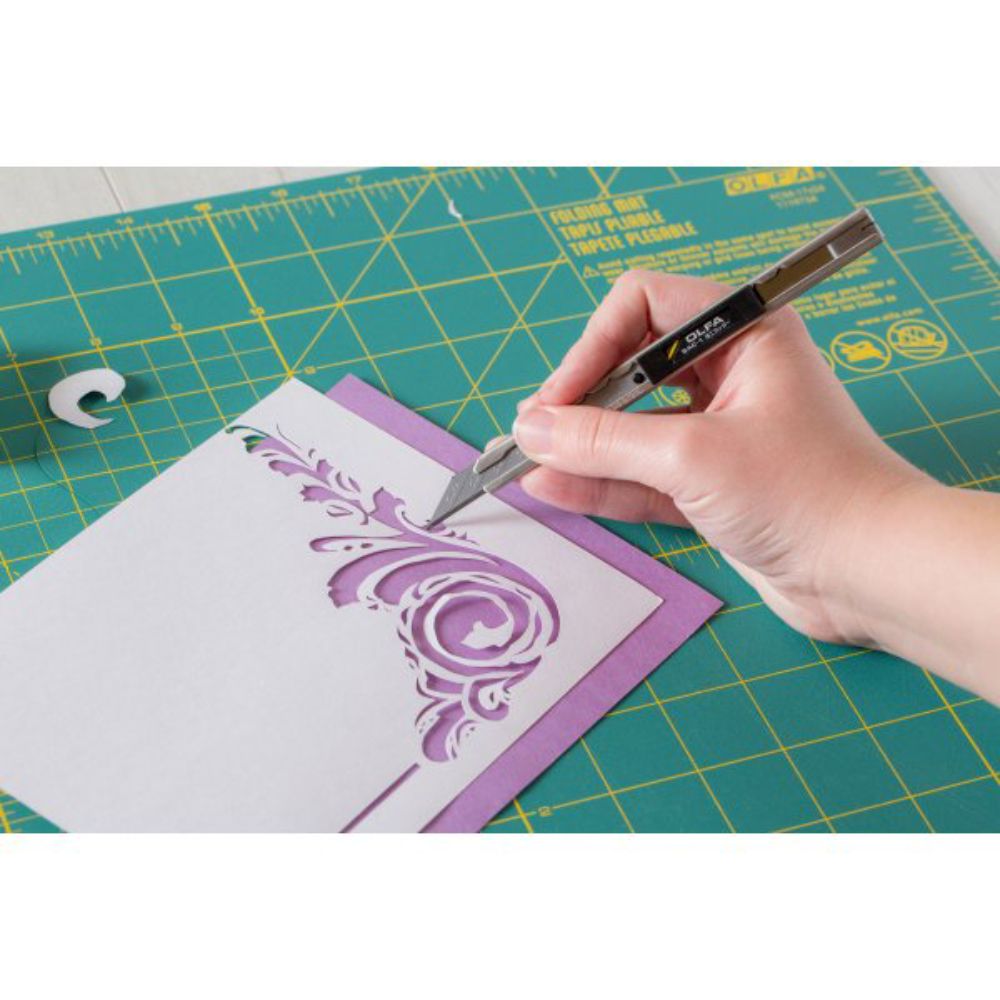 Person using an OLFA precision utility knife to cut an intricate design on a green cutting mat