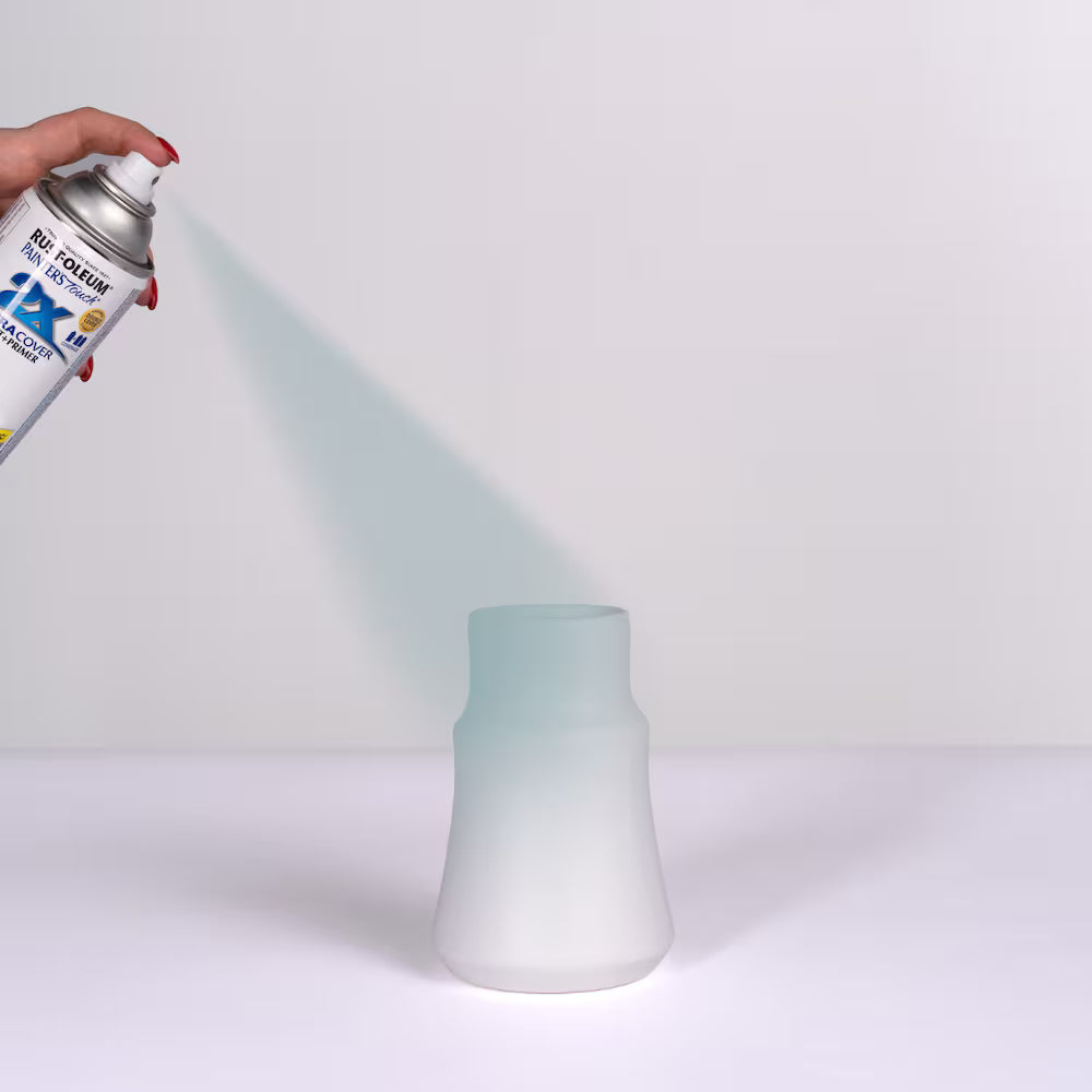 Close-up of a hand using a Rust-Oleum Painter's Touch spray can to apply a smooth coat of grey primer onto a white ceramic vase.