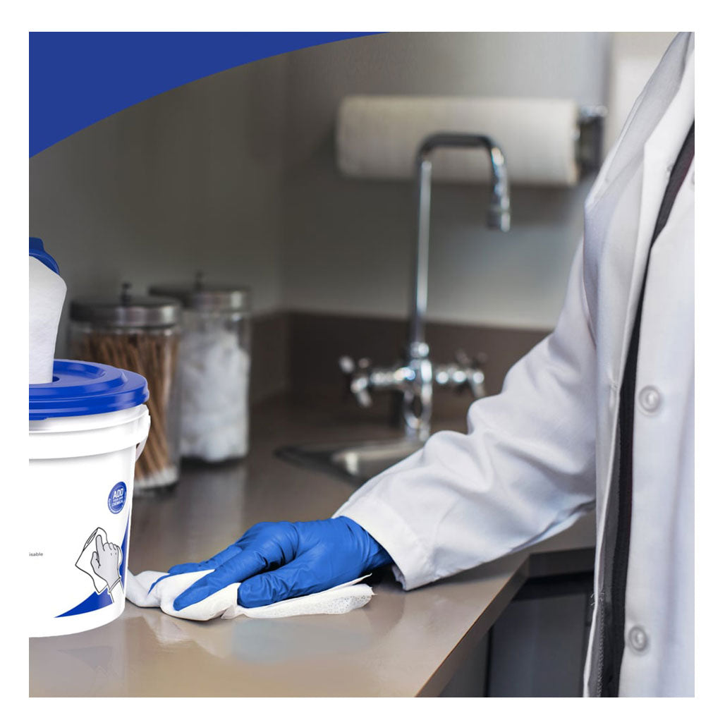 A scientist in a white lab coat and blue nitrile gloves using a white WypAll WetTask critical clean wiper to sanitize a laboratory counter next to a dispensing bucket