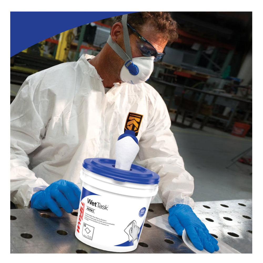 An industrial worker wearing a KleenGuard white protective suit, safety glasses, a respirator mask, and blue nitrile gloves using a white WypAll WetTask wiper on a metal workstation table